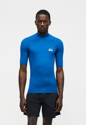 Mees, kes kannab keha järgi lõigatud sinist lühikeste varrukatega ujumispinda, mille rinnal on logo, ja musti ujumispükse, seisab ühevärvilise heleda tausta ees.