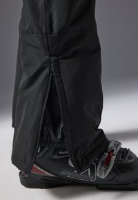 Black ski pants with a side zipper and elastic cuffs. A black ski boot is visible, featuring red accents and a gray buckle.