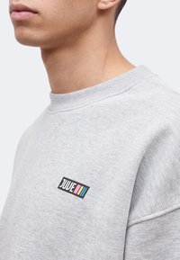 Κοντινό πλάνο ενός ατόμου που φοράει ανοιχτογκρι φούτερ με μικρό ορθογώνιο πολύχρωμο λογότυπο "JUVE" στο στήθος.