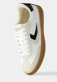 Witte sneaker met zwarte accenten, suède en glad leer materialen, ronde neus, platte witte veters en een gomrubberen buitenzool.