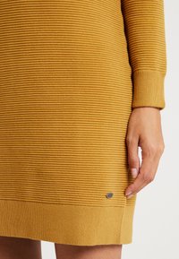 Gros plan sur une robe en maille côtelée jaune moutarde à manches longues, montrant une main reposant à côté de l'ourlet de la robe avec une petite étiquette de marque argentée.