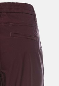 Burgunderfarbene Hose mit glatter Textur, elastischem Bund und einer einzelnen Gesäßtasche. Mit kontrastierenden Nähten und einer taillierten Passform.