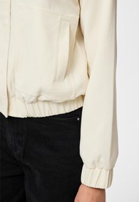 Jakna boje kreme s prednjim džepom i elastičnim manžetnama, nošena s crnim hlačama, vidi se sredina tijela i desna ruka.