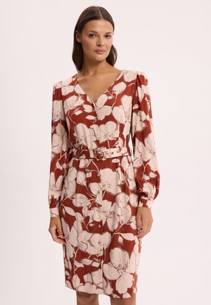 Kobieta ubrana w rdzawobrązową, długą sukienkę z paskiem, długim rękawem, dużymi białymi kwiatowymi wzorami i guzikami z przodu na jednolitym tle.