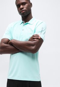 Άνδρας με γαλάζιο πόλο μπλουζάκι Calvin Klein με κουμπιά, σταυρωμένα χέρια, εμφανιζόμενος από τη μέση και πάνω σε απλό φόντο.