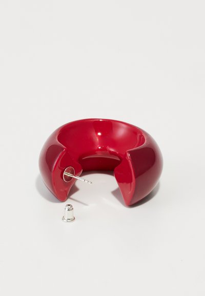 Roter glänzender Armreif mit Splittdesign, ausgestattet mit einem Metallstift, liegt neben einer silbernen Kappe. Glatte Oberfläche mit einer geschwungenen, klobigen Form.