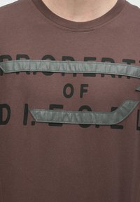 Μπορντό βαμβακερό T-shirt με μαύρο έντονο κείμενο και υφασμάτινες γκρι υφές στο στήθος, με σχέδιο λαιμόκοψης.