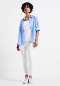 Chemise en lin bleu clair, débardeur blanc, leggings blancs et baskets blanches. Cheveux bouclés, posture debout, accessoires minimes.