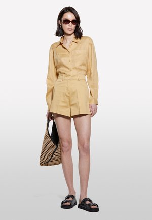 Chemise en lin jaune à manches longues, short plissé jaune assorti, sandales noires et sac tissé. Design épuré et minimaliste.