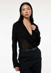 Blusa nera con scollo drappeggiato, tessuto trasparente, maniche lunghe e polsini con bottone. Abbinata a pantaloni scuri su misura e una cintura nera.