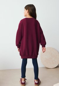 Una ragazza in un maglione oversize granata e leggings blu navy sta in piedi con le spalle al pubblico, di fronte a uno sfondo semplice e chiaro.
