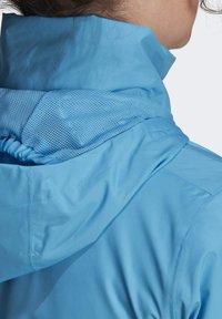Lichtblauwe jas met hoge kraag en mesh-voering in de kap, voorzien van een gladde stoftextuur en geplooide details aan de rand van de kap.