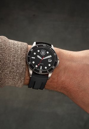 Wrist with black dive watch featuring round white markers, rotating bezel, date window, silver case, black rubber strap, and beige sweater sleeve.