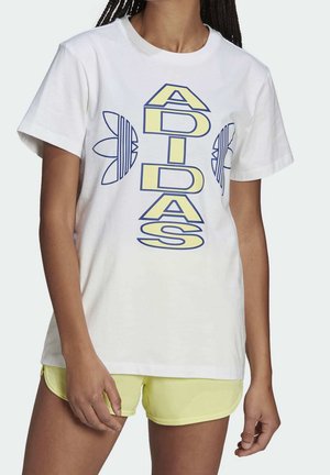 Kobieta ubrana w białą koszulkę Adidas z pionowym żółtym logo i niebieskimi wzorami koniczyny, połączona z jasnżółtymi szortami.