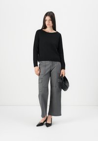 Pull noir à manches longues, associé à un pantalon cargo gris avec de multiples poches. Le modèle tient un sac noir. Talons aiguilles noirs.