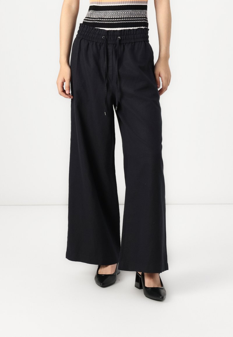 Schwarzweite Baumwollhosen mit einem elastischen Bund und Kordelzug. Glatte Textur und knöchellanges Design, kombiniert mit schwarzen Absätzen.