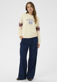 Sudadera crema con acentos a rayas en azul marino y burdeos, que presenta un gráfico "CLÁSICO 86". Pantalones vaqueros de pierna ancha en denim oscuro con pliegues.