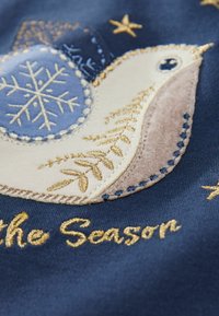 Broderet stof med et fuglemotiv, snefnug, gyldne detaljer og teksten "Fejr sæsonen" på en mørkeblå baggrund.
