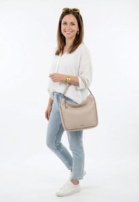 Donna sorridente con indosso una camicetta bianca, jeans azzurri chiari, sneakers bianche, occhiali da sole gialli sulla testa, orologio d'oro, che porta una borsa beige su sfondo bianco.