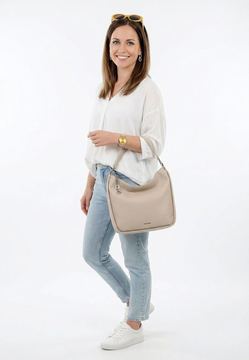 Donna sorridente con indosso una camicetta bianca, jeans azzurri chiari, sneakers bianche, occhiali da sole gialli sulla testa, orologio d'oro, che porta una borsa beige su sfondo bianco.