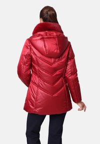 Chaqueta acolchada roja con capucha, cuello de piel sintética y patrón de acolchado en forma de chevron. Textura suave con un acabado brillante.