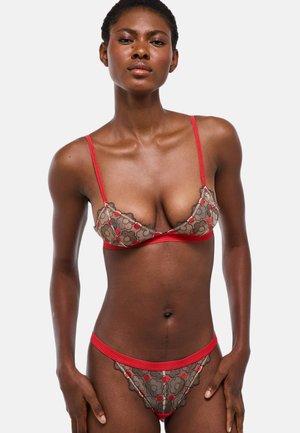 Femme portant un soutien-gorge en dentelle florale rouge et beige assorti à sa lingerie, debout devant un fond uni gris clair.