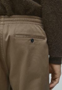 Brązowe spodnie materiałowe o gładkiej fakturze, wyposażone w tylną kieszeń z zapięciem na guzik oraz elastyczną talię.