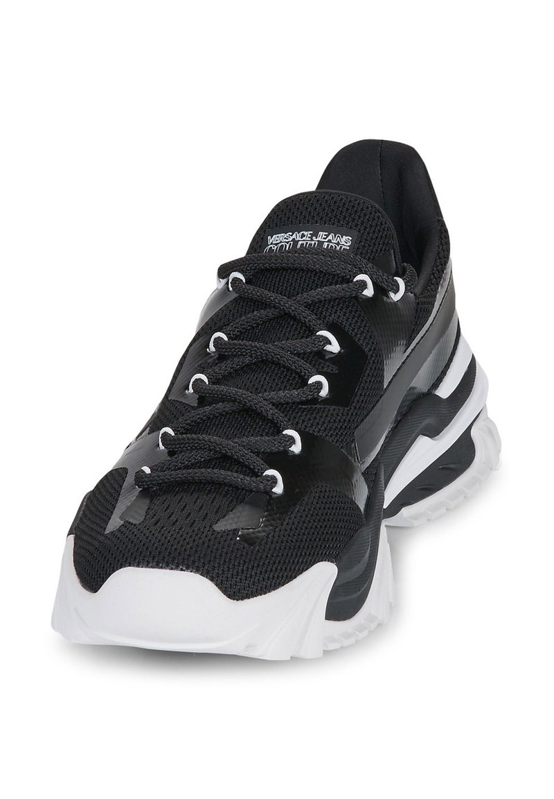 Zapato deportivo negro con parte superior de malla, acentos brillantes en contraste, suela blanca y sistema de cordones distintivo. Presenta el logo de la marca en la lengüeta.