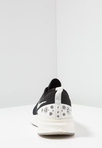 Černá atletická obuv s texturovaným svrškem, bílou patou zdobenou květinovými vzory a pruhovaným poutkem. Gumová podešev s viditelným polstrováním.