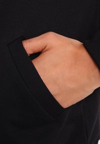 Hand gedeeltelijk in de voorkant van een zwart lang mouwenshirt gestoken, wat een contrast laat zien tussen de huidkleur en de stof.