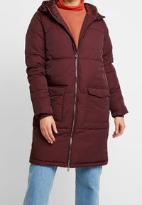 Giacca puffer borgogna con cappuccio, zip frontale e due tasche sul petto; presenta un design trapuntato e una vestibilità comoda, abbinata a jeans azzurri chiari.