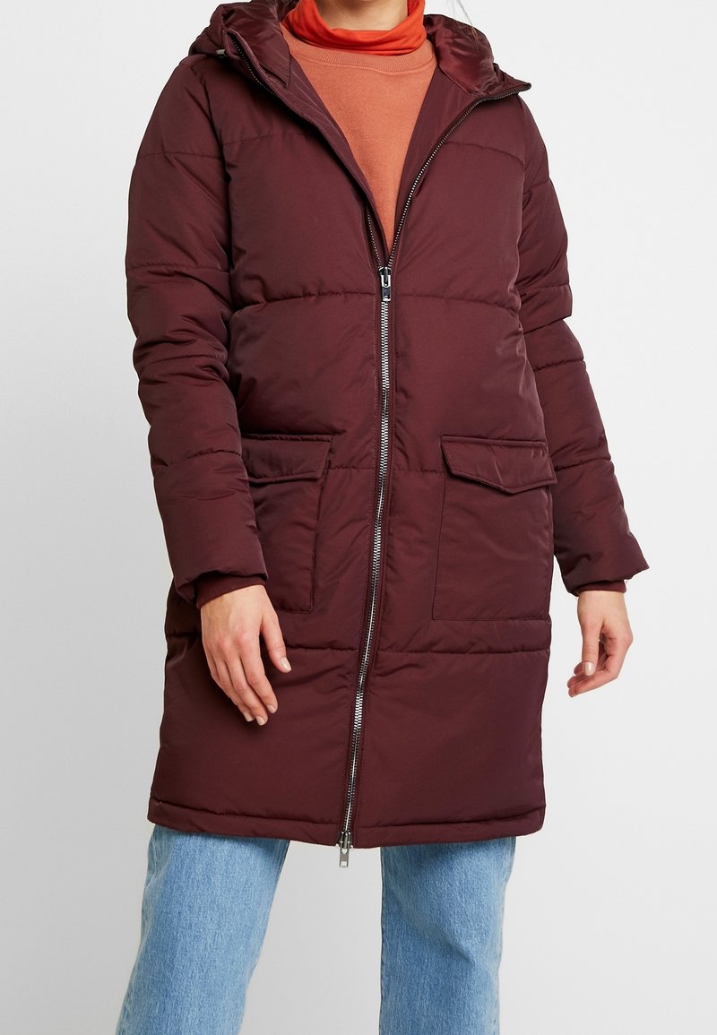 Giacca puffer borgogna con cappuccio, zip frontale e due tasche sul petto; presenta un design trapuntato e una vestibilità comoda, abbinata a jeans azzurri chiari.