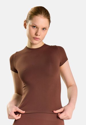 Ung kvinde med lys hud og blå øjne iført en tætsiddende brun kortærmet top, der holder i bundkanten med begge hænder.