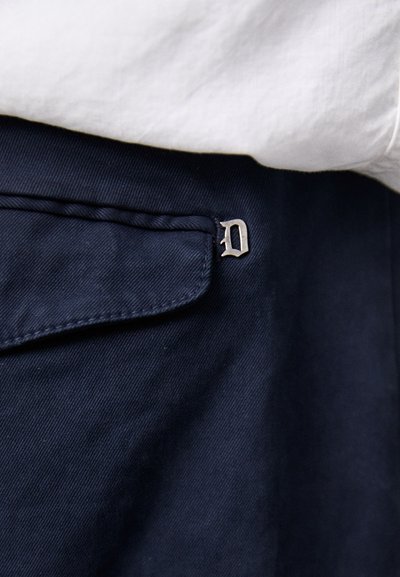 Los pantalones azul marino tienen una textura suave, costuras sutiles y un acento de logo metálico en forma de la letra "D" en el bolsillo frontal.