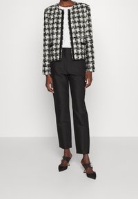 Personne portant une veste à carreaux noir et blanc, une chemise blanche, un pantalon droit noir et des talons pointus noirs ornés de décorations.