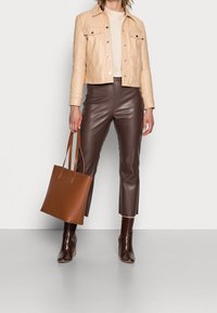 Veste en cuir beige, haut crème, pantalon marron en similicuir et sac fourre-tout marron. Bottines marron à talons avec une texture croco.