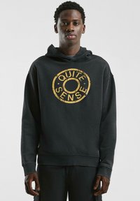 Schwarzer Kapuzenpullover aus weichem Stoff, mit einem abgenutzten goldenen runden Logo mit dem Text "QUITE SENSE" in der Mitte.
