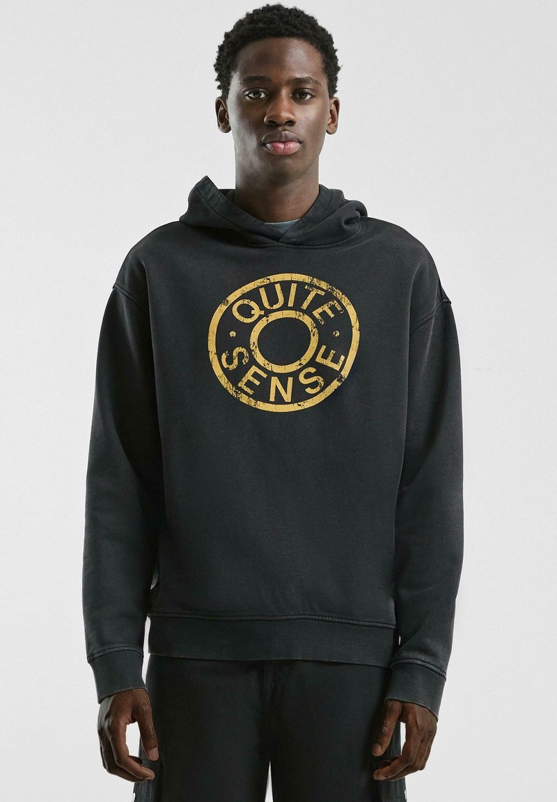 Schwarzer Kapuzenpullover aus weichem Stoff, mit einem abgenutzten goldenen runden Logo mit dem Text "QUITE SENSE" in der Mitte.