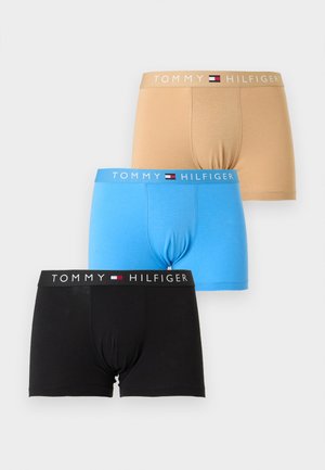 Młody mężczyzna stoi boso, ubrany w czarne bokserki Tommy Hilfiger, lekko patrzy w lewo na jednolitym jasnym tle.