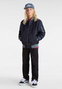 Chaqueta bomber azul con acentos grises, combinada con una camisa roja y pantalones negros. La persona lleva una gorra gris y zapatillas negras.