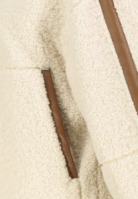 Cremfarbener Fleece-Jacke mit strukturiertem Oberflächen, mit braunen Lederakzenten entlang der Nähte und Taschen.
