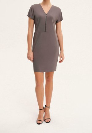 Femme portant une robe taupe courte à manches courtes, longueur genou, avec fermeture éclair devant, associée à des escarpins noirs ouverts à bride cheville, debout sur fond blanc.