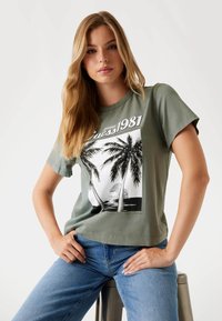 Maslinasto zelena pamučna majica s grafičkim printom koji prikazuje palme, plažu i natpis "Guess 1981", okrugli izrez, kratki rukavi.