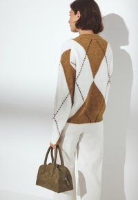 Pull en tricot à motifs en losanges marron et blanc, texture en maille, associé à un sac à main en suède vert olive, tenu par une personne portant un pantalon blanc.