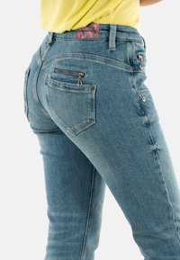 Vaqueros de denim azul descolorido con un diseño ajustado, que cuentan con dos bolsillos con cremallera y una etiqueta con un patrón floral en la cintura.