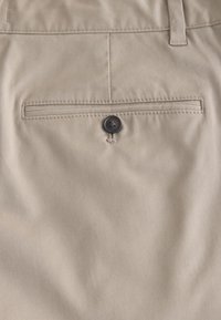 Beige tyg i närbild som visar en bakficka med en horisontell slits och en mörk knappstängning på skräddarsydda byxor.