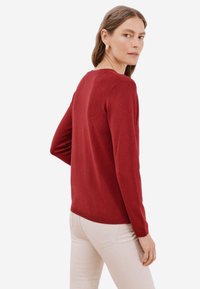 Maglione rosso lavorato a maglia con maniche lunghe, scollatura tonda e silhouette aderente. Il materiale appare morbido, con una texture liscia.