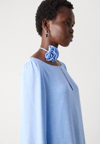 Lichtblauw blouse met witte stippen, vloeiende mouwen, sleutelgat halslijn; gecombineerd met een blauwe bloemen choker en grote ronde oorbellen.