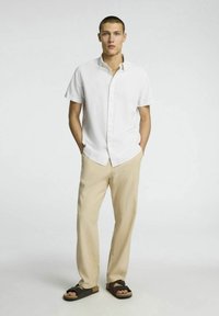 Vit kortärmad skjorta med knappar, beige tapered-byxor och bruna sandaler med korksål. Modellen står med händerna i fickorna.