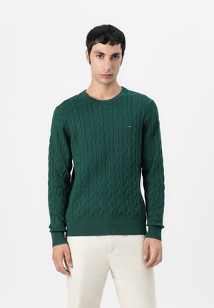 Grønn kabelstrikket genser med rund hals, ribbestrikkede ermer og kant, med en liten logoaksent på brystet. Modellen har på seg lyse bukser.